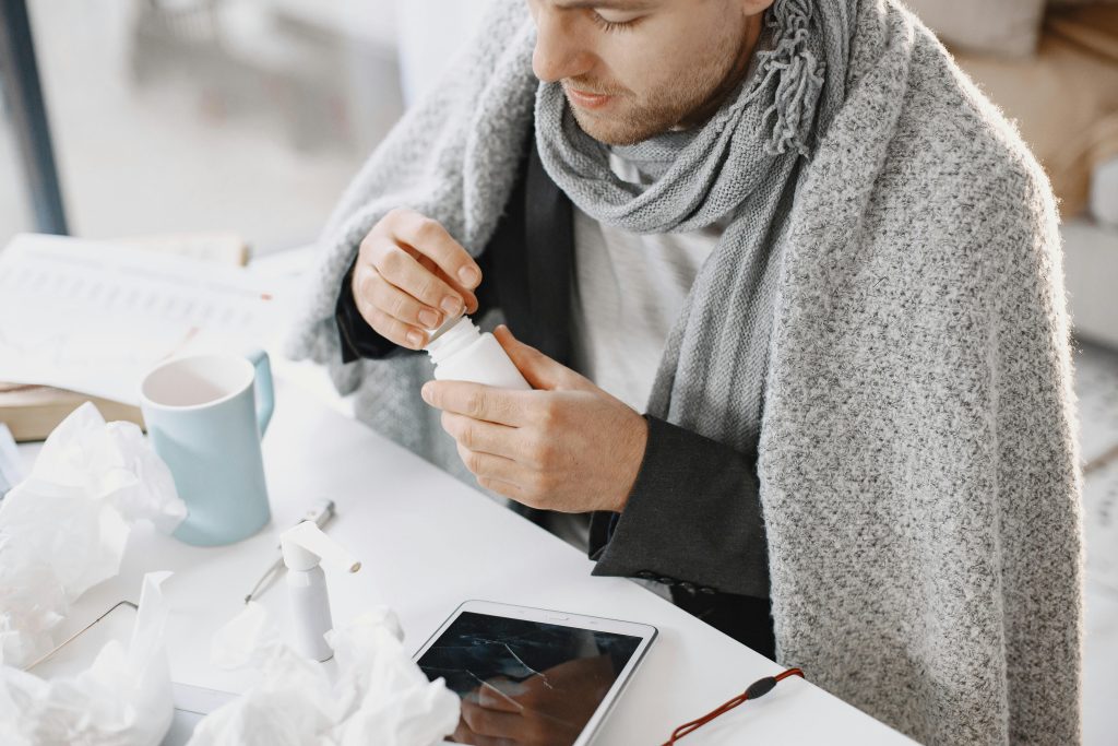 Adult male in scarf taking medicine at a desk with tissues and tablet, feeling unwell.